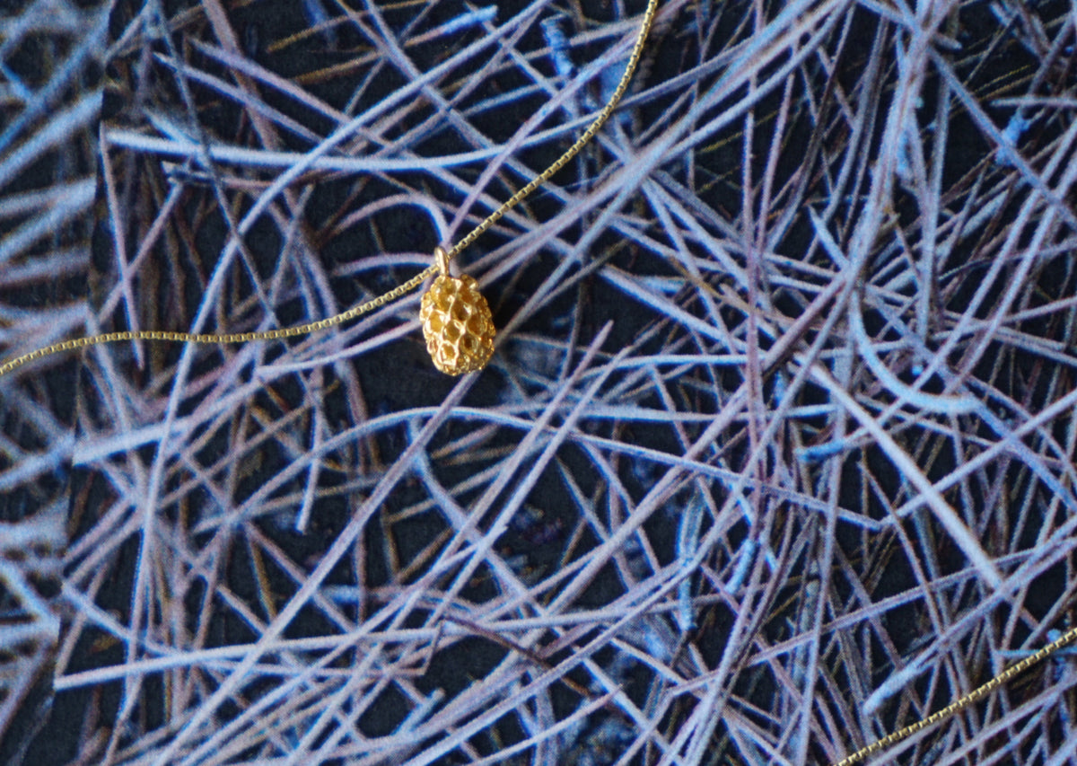 A gold pinecone-shaped pendant necklace on a delicate gold chain, set against a dark background of intertwined blue and white fibers.