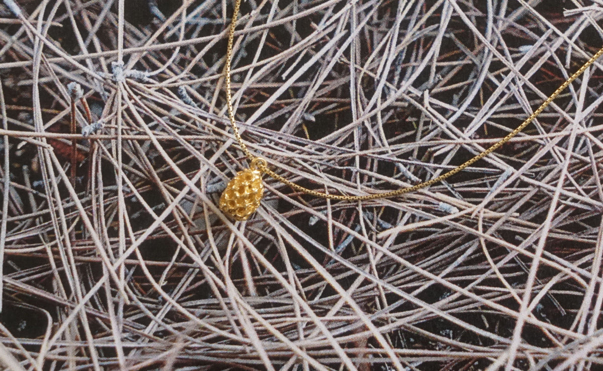 A gold pinecone-shaped pendant necklace resting on a bed of dry brown pine needles outdoors.